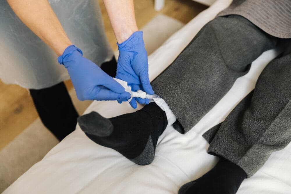 A caregiver in blue gloves adjusts a medical device on a patient's ankle who is lying on a bed. - Home Instead