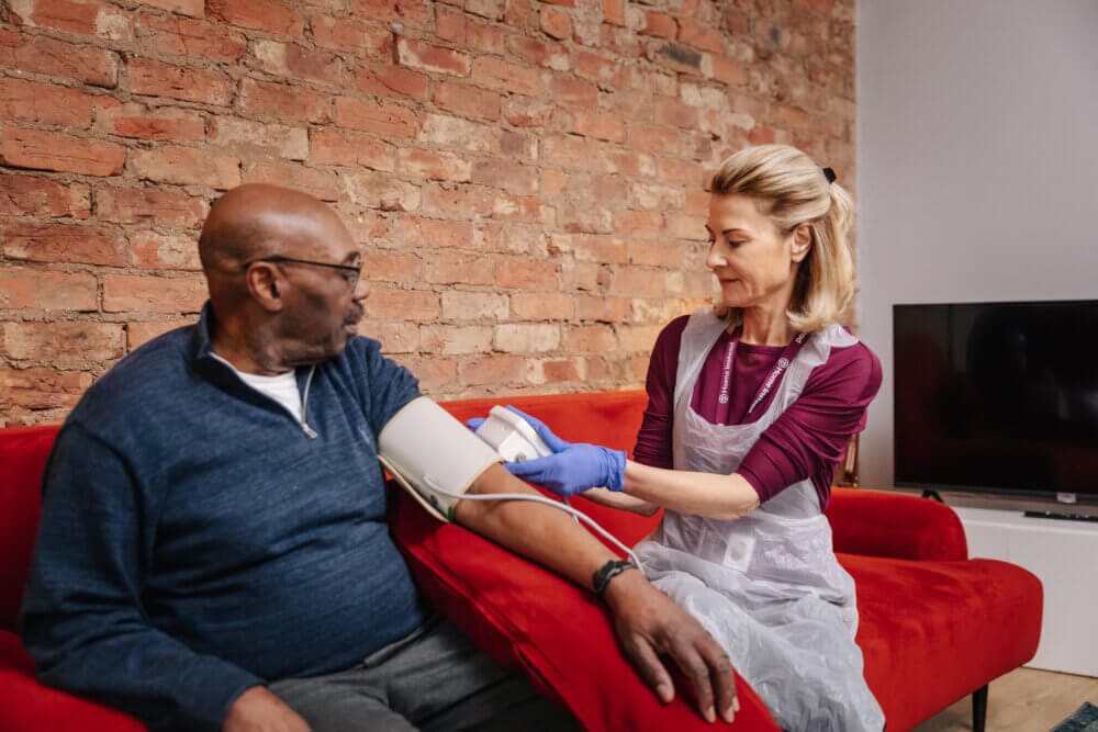A healthcare worker checks the blood pressure of a man sitting on a red couch, with a brick wall in the background. - Home Instead