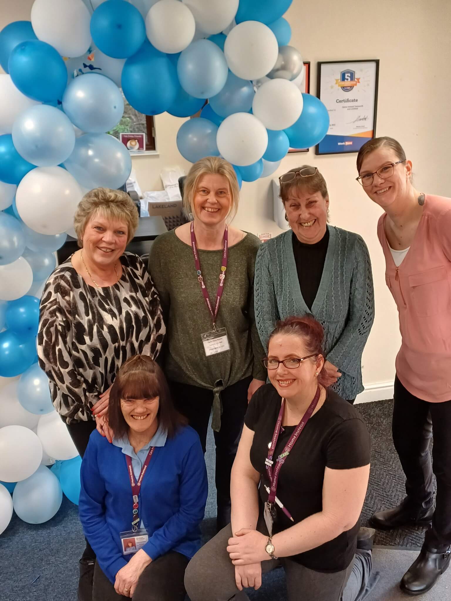 Six care professionals and smiling, with a balloon arch in the background and a certificate on the wall behind them. - Home Instead