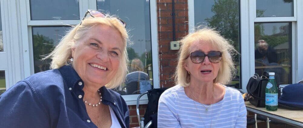 Two older women with sunglasses, one smiling, the other with a neutral expression, sit outside near a brick building. - Home Instead