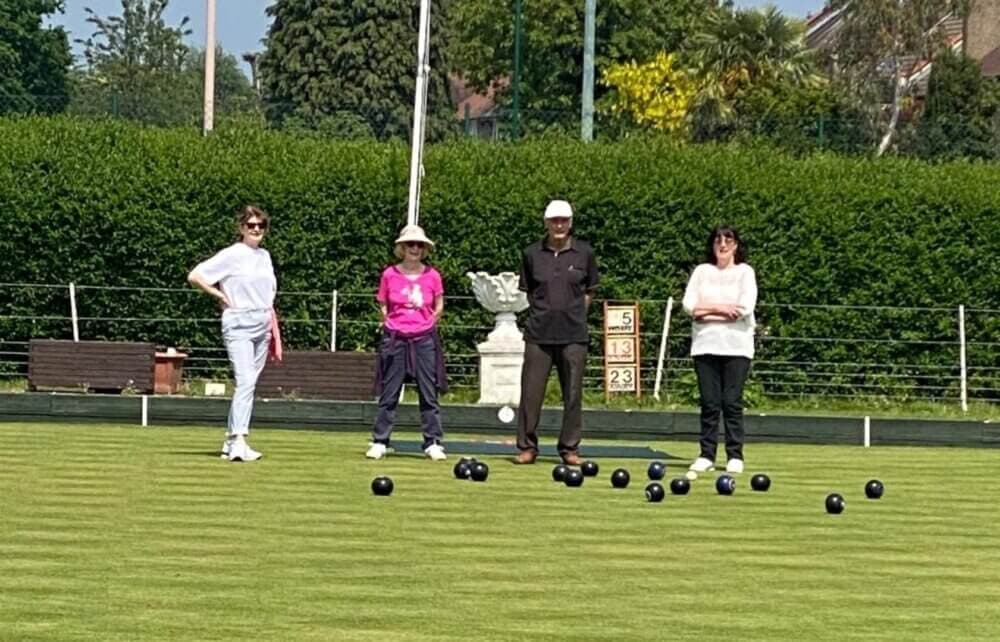 Four people standing on a lawn bowling green, with bowls scattered on the grass. Trees and a hedge in the background. - Home Instead