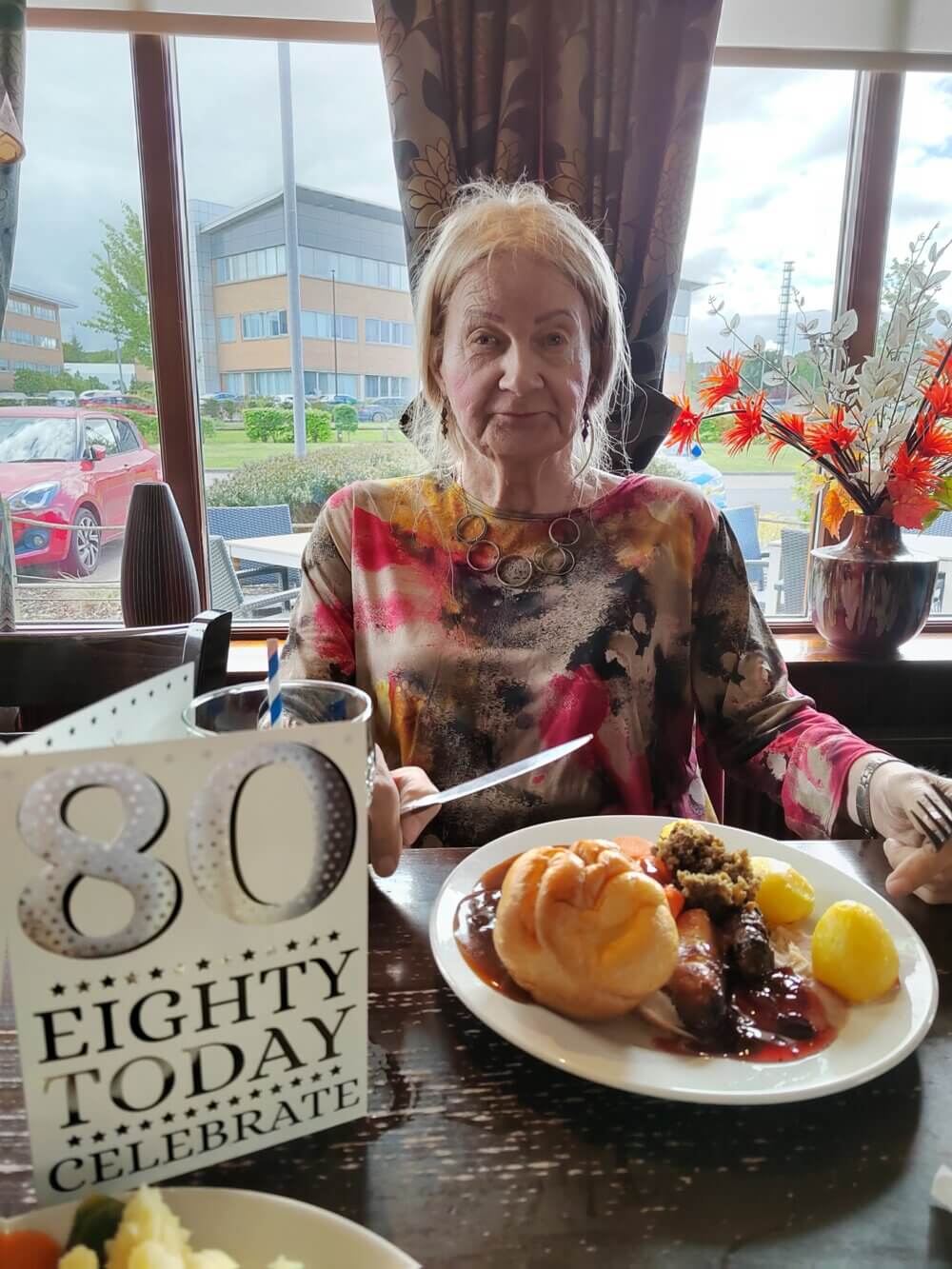 Elderly woman celebrating her 80th birthday with a meal at a restaurant, sign on table reading "80 Eighty Today Celebrate. - Home Instead