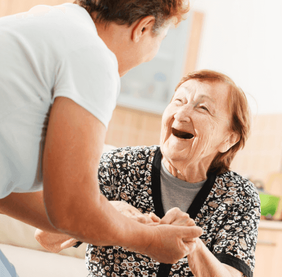 Elderly woman smiling and holding hands with a caregiver in a bright room. - Home Instead