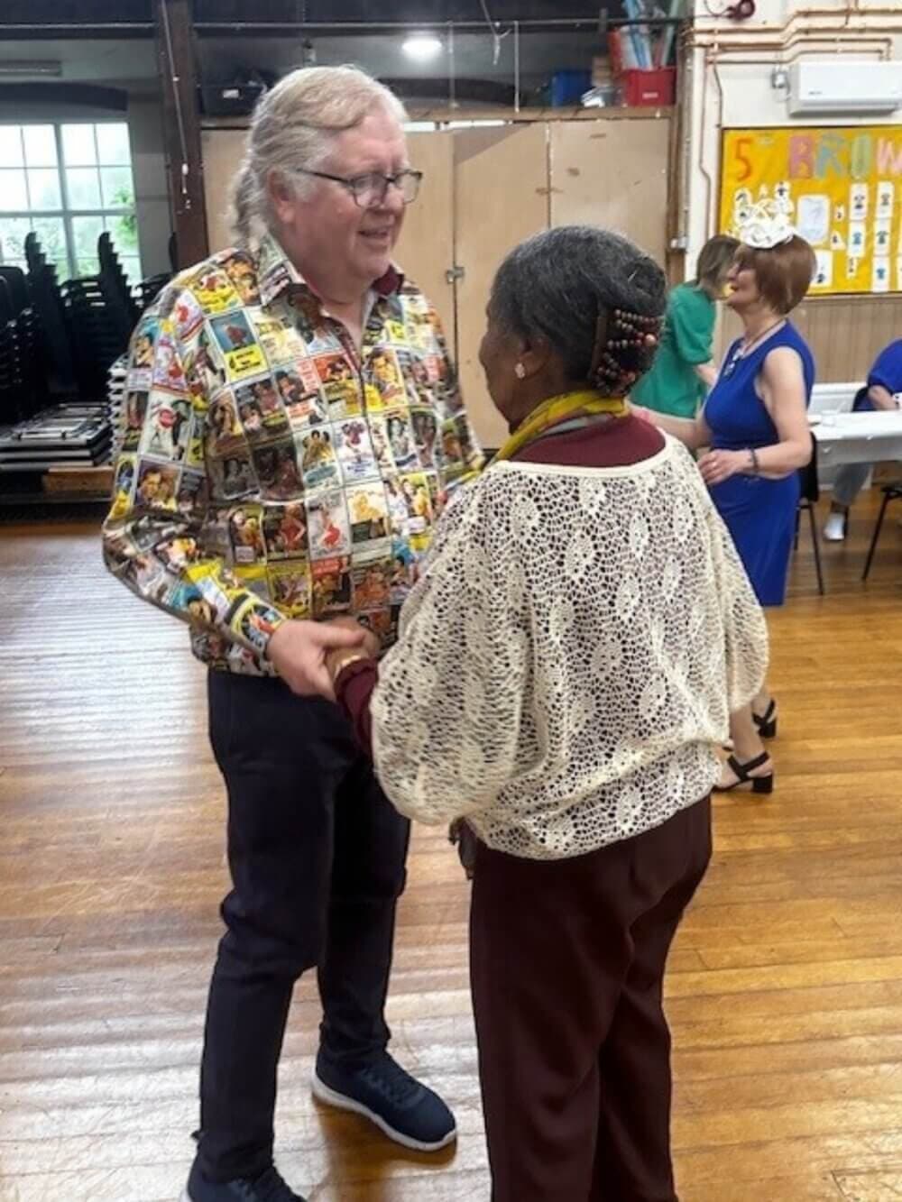 Two people holding hands, engaged in conversation on a wooden floor in a well-lit room with other people in the background. - Home Instead