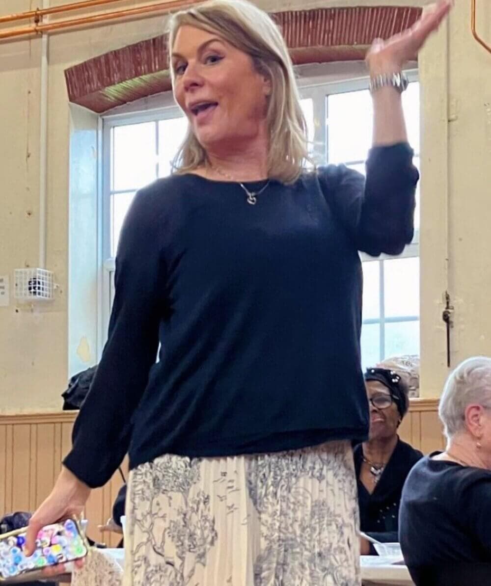 A woman holding a phone gestures with her hand while standing in front of a window with people seated behind her. - Home Instead