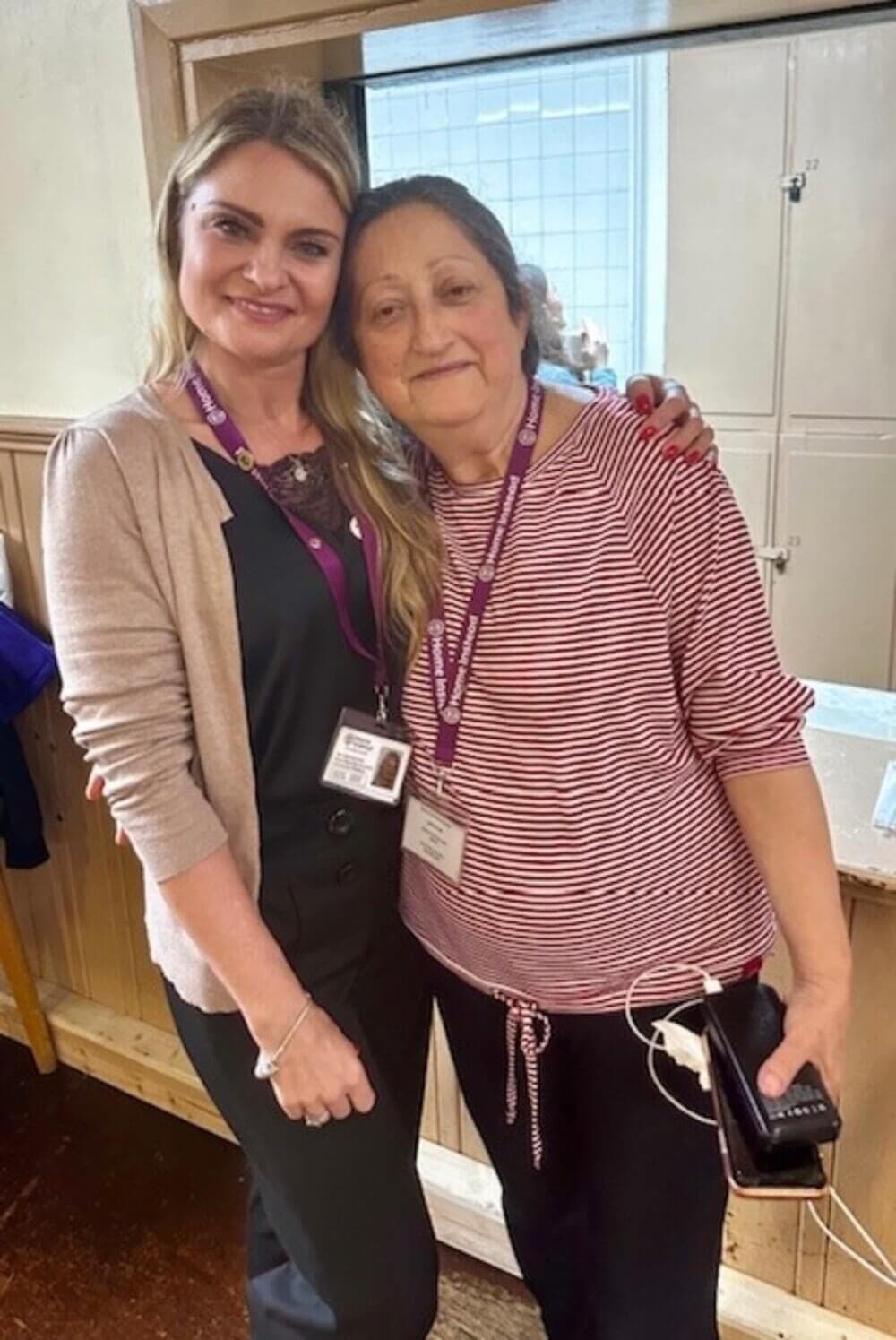 Two women standing together, smiling. Both are wearing lanyards and ID badges. One is holding a phone with attached earphones. - Home Instead