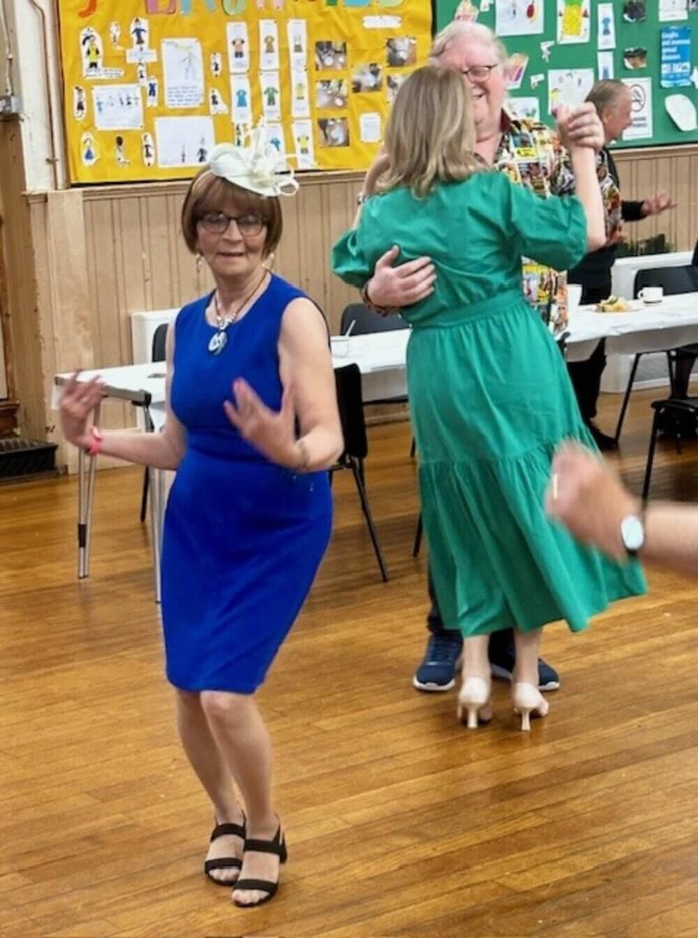 People dancing in a community hall, with a woman in a blue dress in the foreground and a couple dancing in the background. - Home Instead