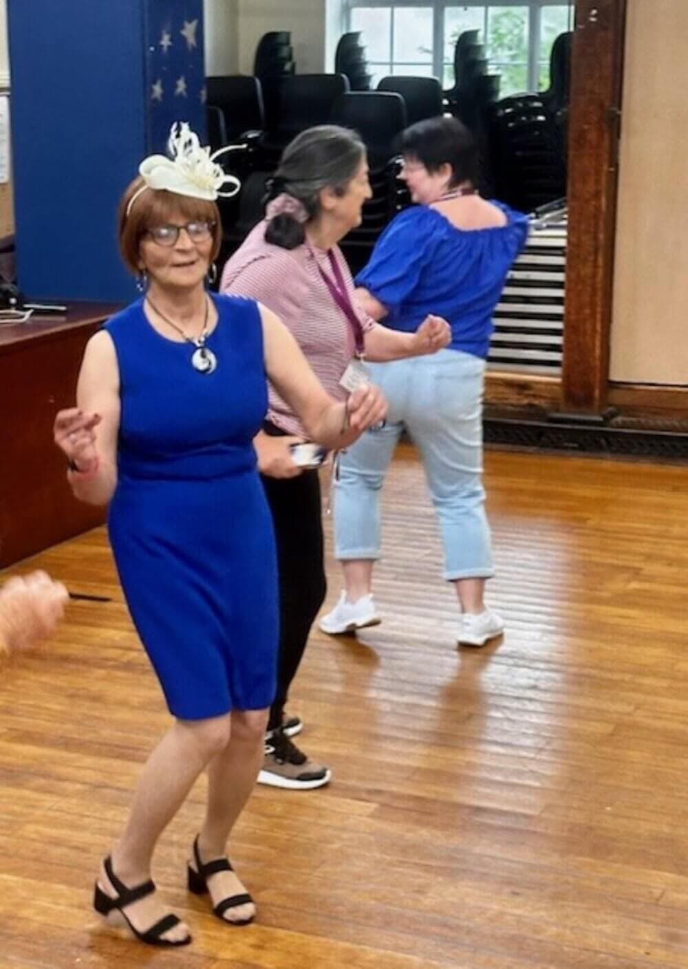 Four women dance in a hall, one in a blue dress and white hat, others in casual attire. Chairs are stacked in the background. - Home Instead