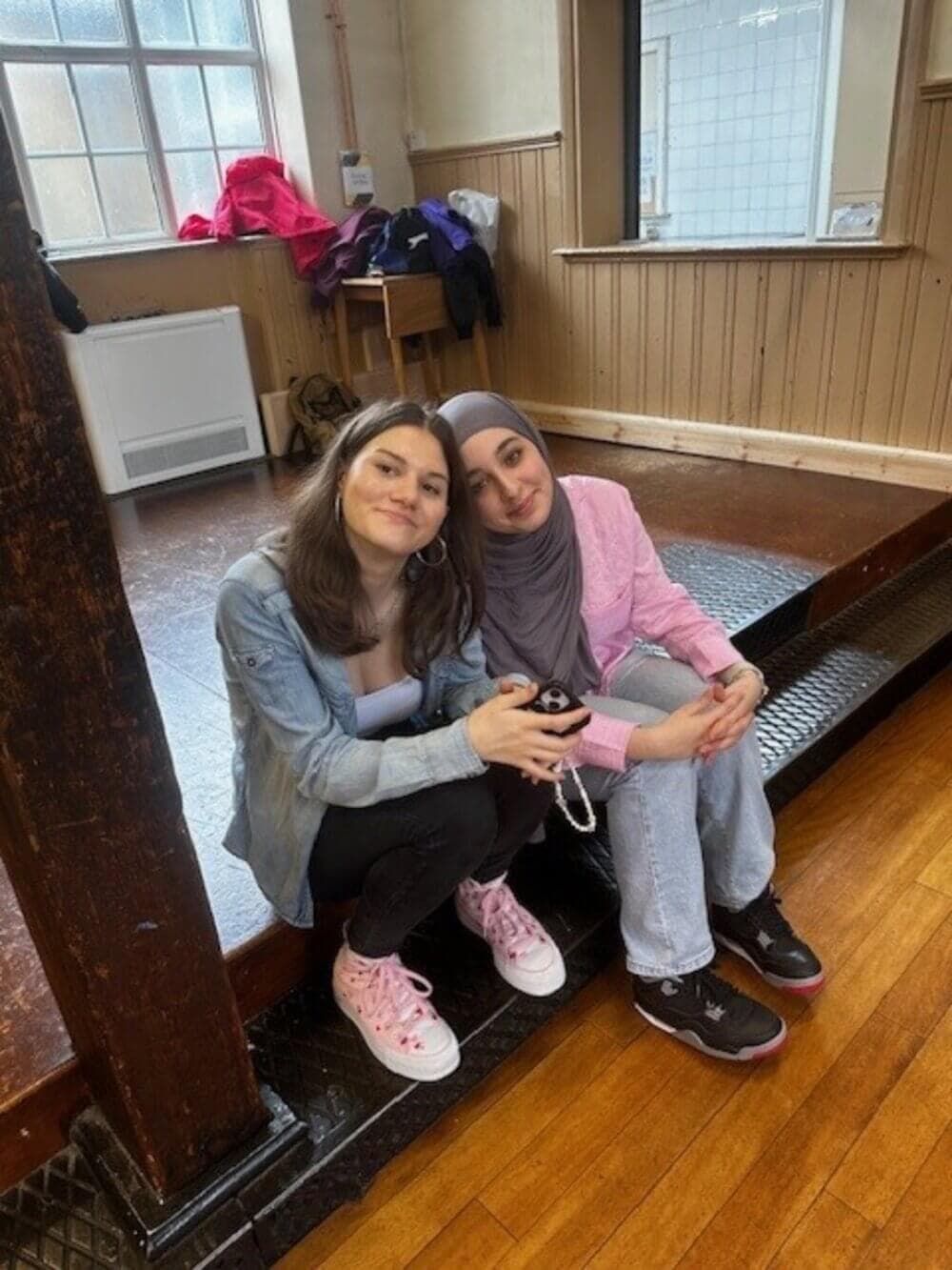 Two women seated on a wooden stage, smiling and leaning towards each other in a bright room with coats hanging in the background. - Home Instead