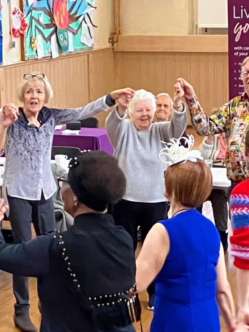 A group of people dance in a circle at a social event, smiling and holding hands. - Home Instead