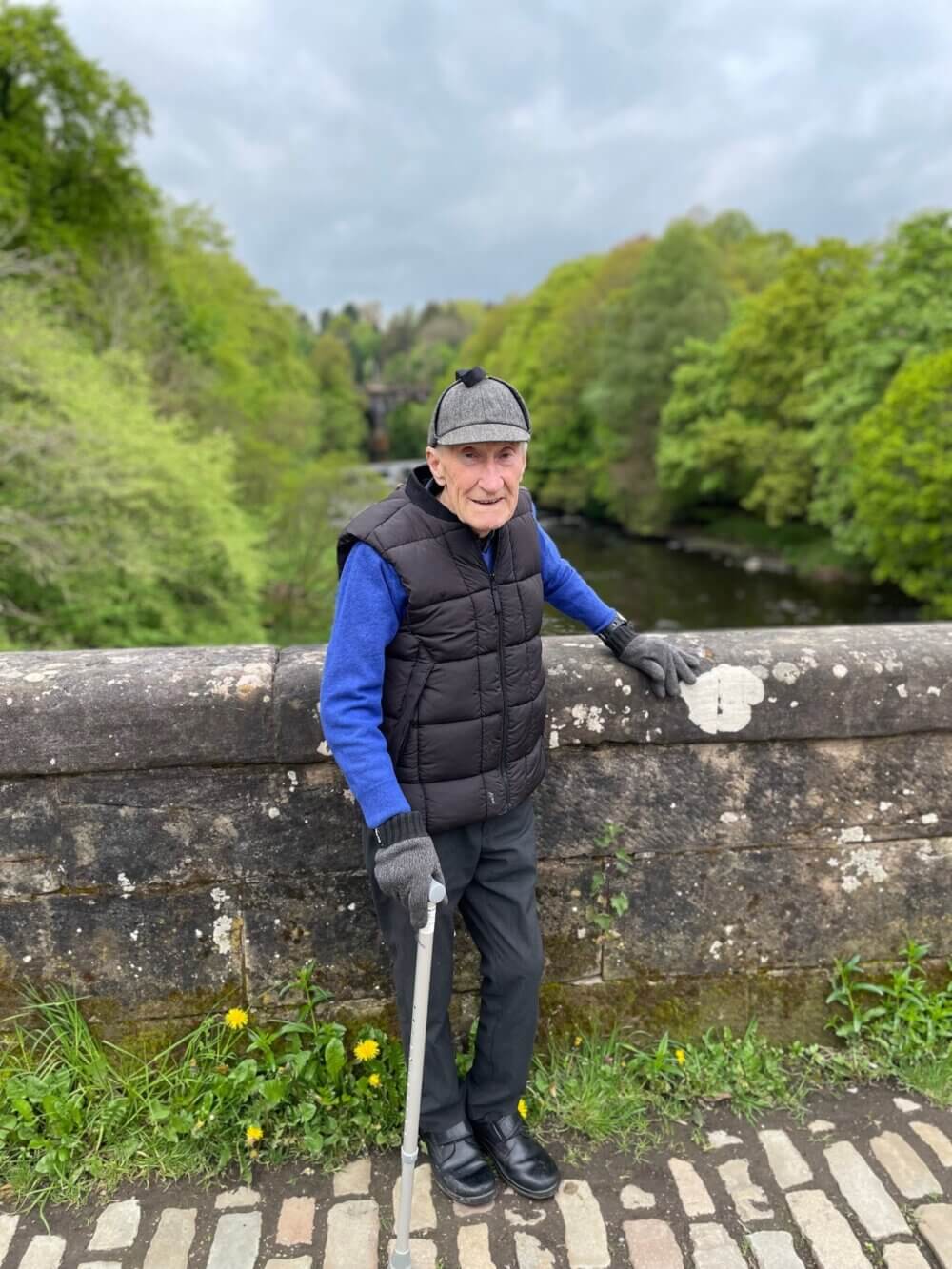 Elderly man with a cane, wearing a hat and vest, stands on a stone bridge with a river and trees in the background. - Home Instead