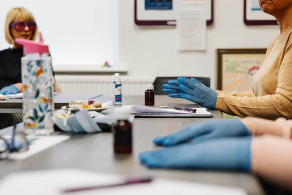 People wearing gloves sitting at a table with various small bottles, papers, and stationery items. - Home Instead