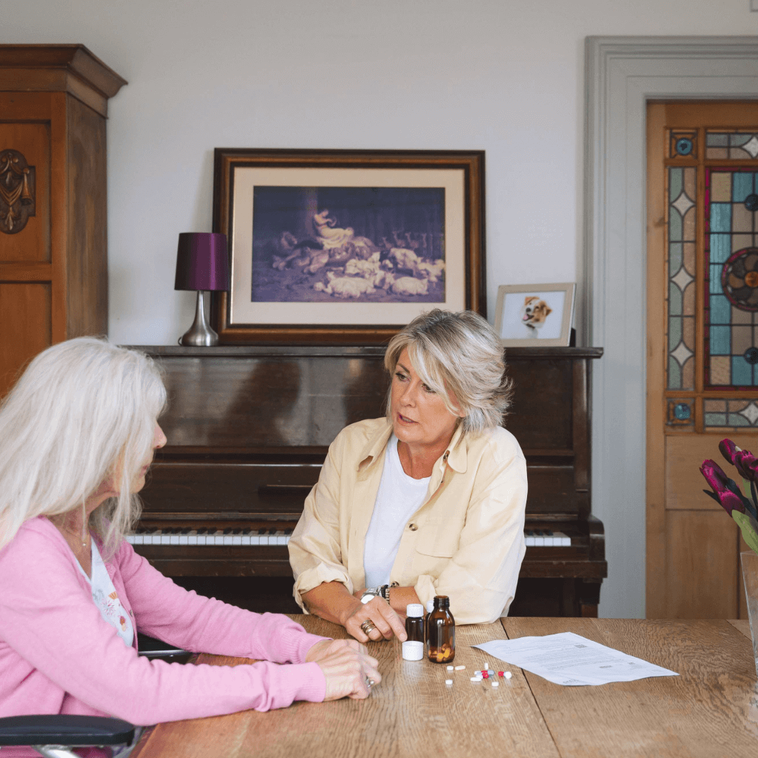 Two women sitting at a table with medications, having a conversation in a cozy room with a piano and artwork. - Home Instead