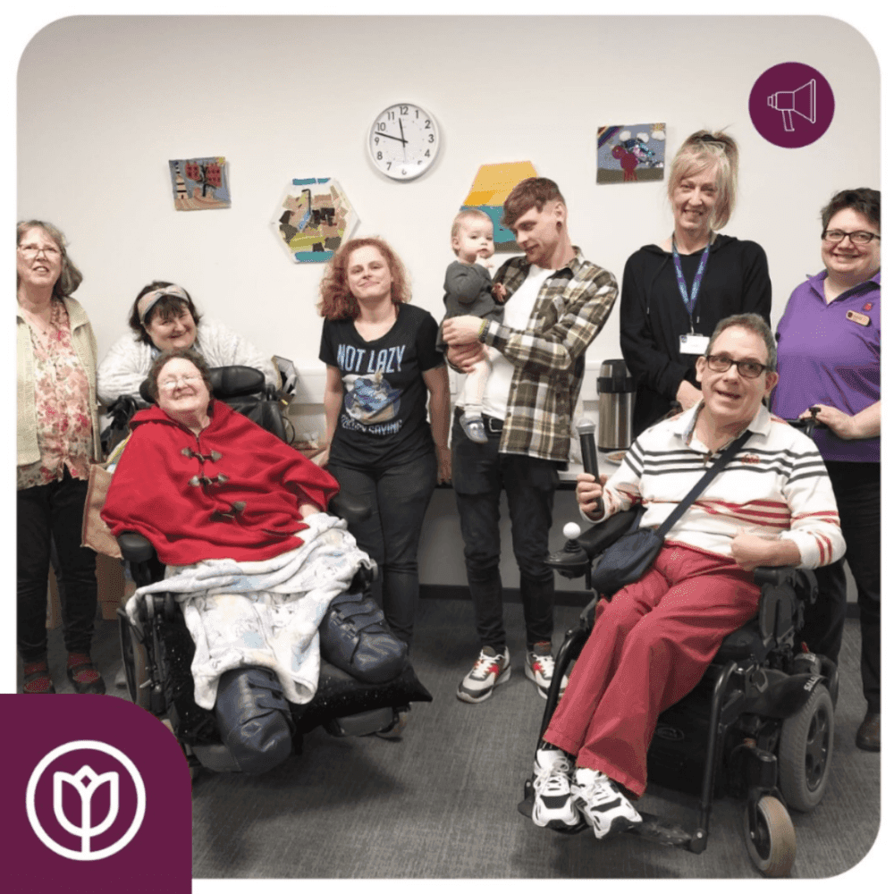 A group of people, some in wheelchairs, smiling and posing indoors in front of a wall with a clock and artwork. - Home Instead
