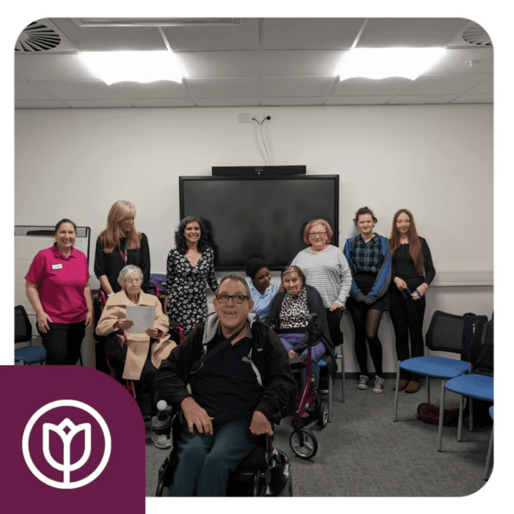 Group of people, some seated and some standing, smiling in a well-lit room with a TV screen on the wall. - Home Instead