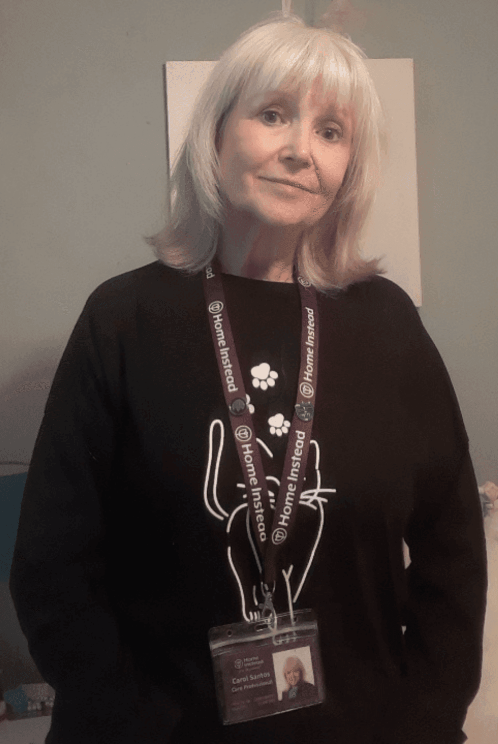 Person with blond hair in a black shirt, wearing a "Home Instead" lanyard and ID badge, standing against a light blue wall. - Home Instead