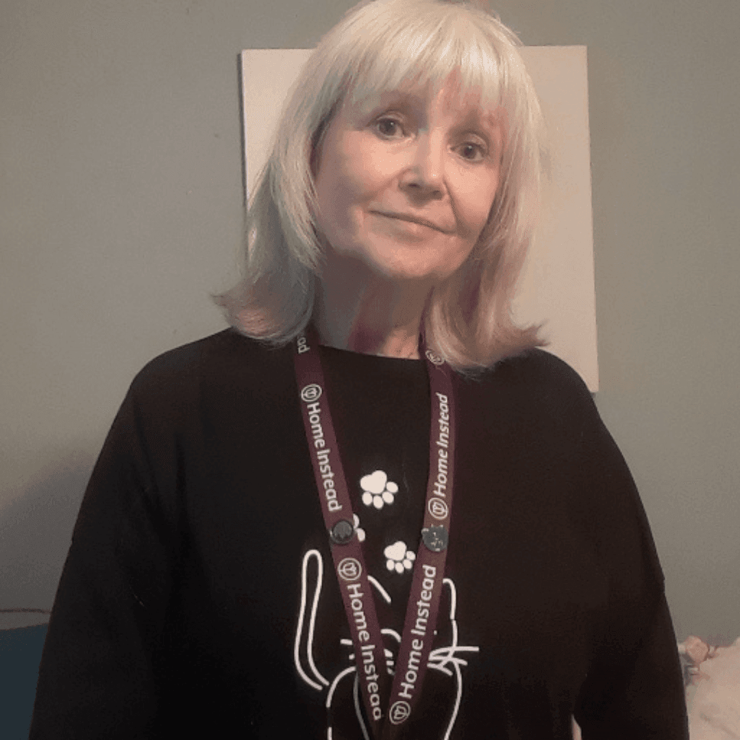 Woman with blond hair wearing a black shirt and Home Instead lanyard standing against a blue wall. - Home Instead