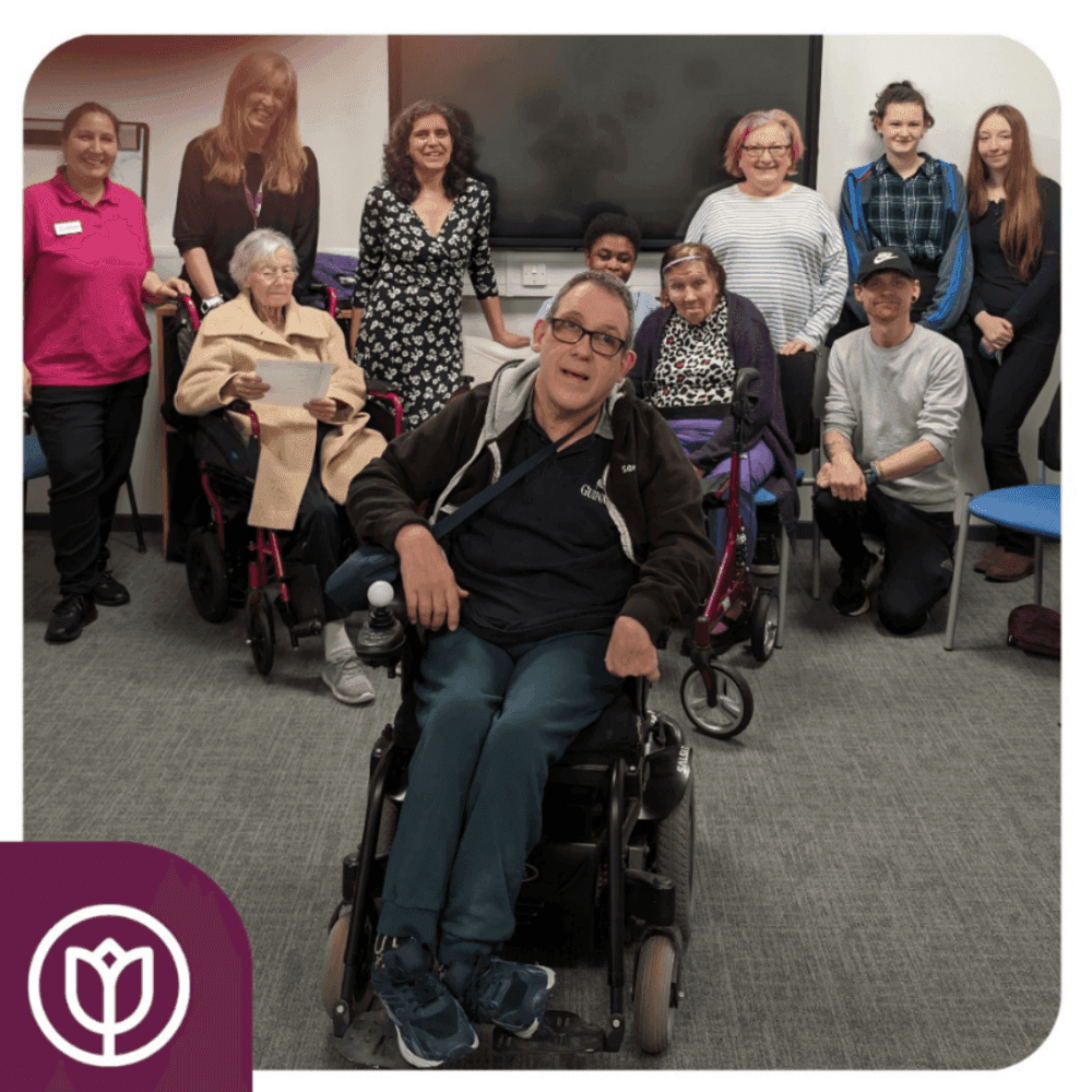 Group of people, including some in wheelchairs, smiling in a room. A logo is in the bottom left corner. - Home Instead