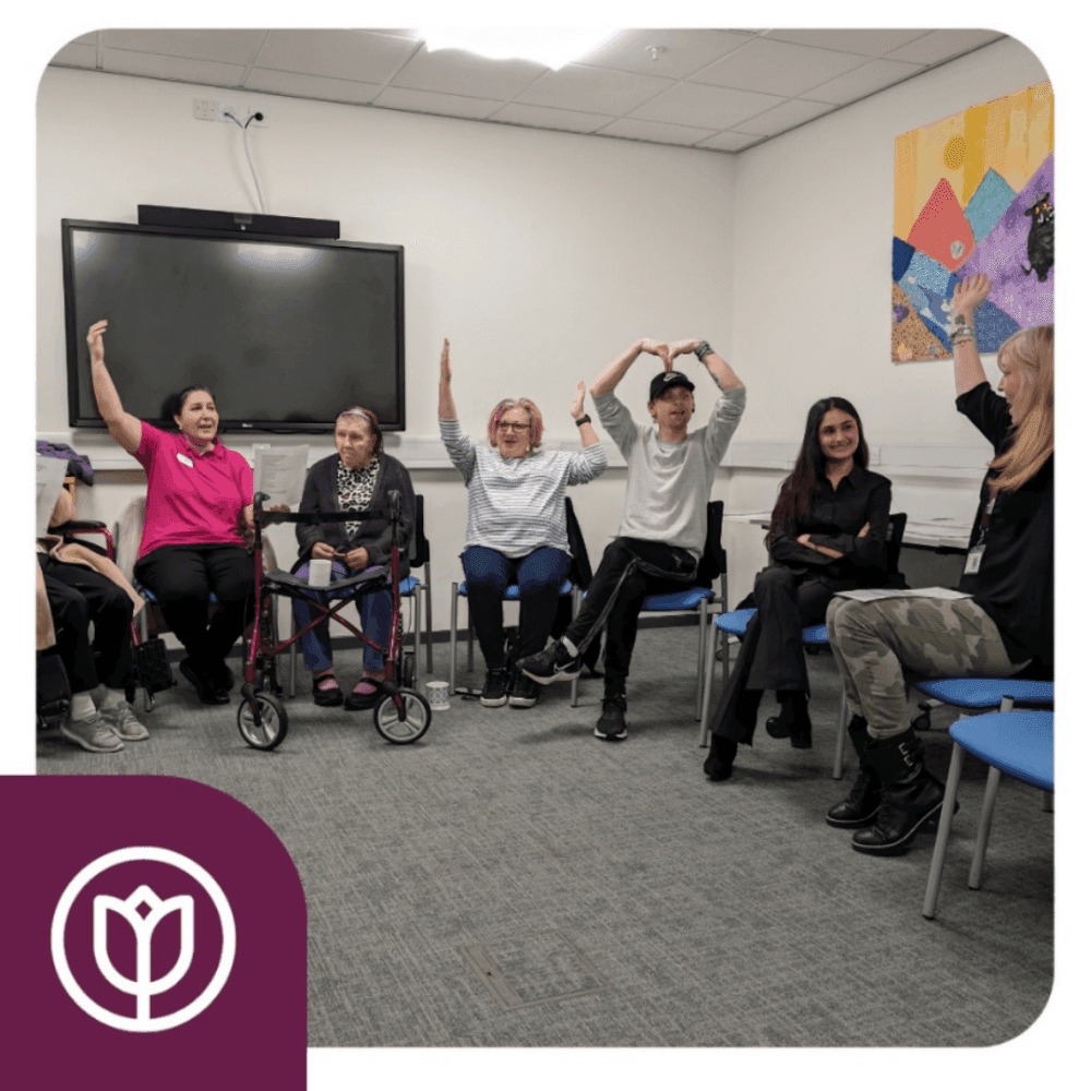 A group of people sitting in a circle in a room, some with raised arms, participating in an activity. - Home Instead