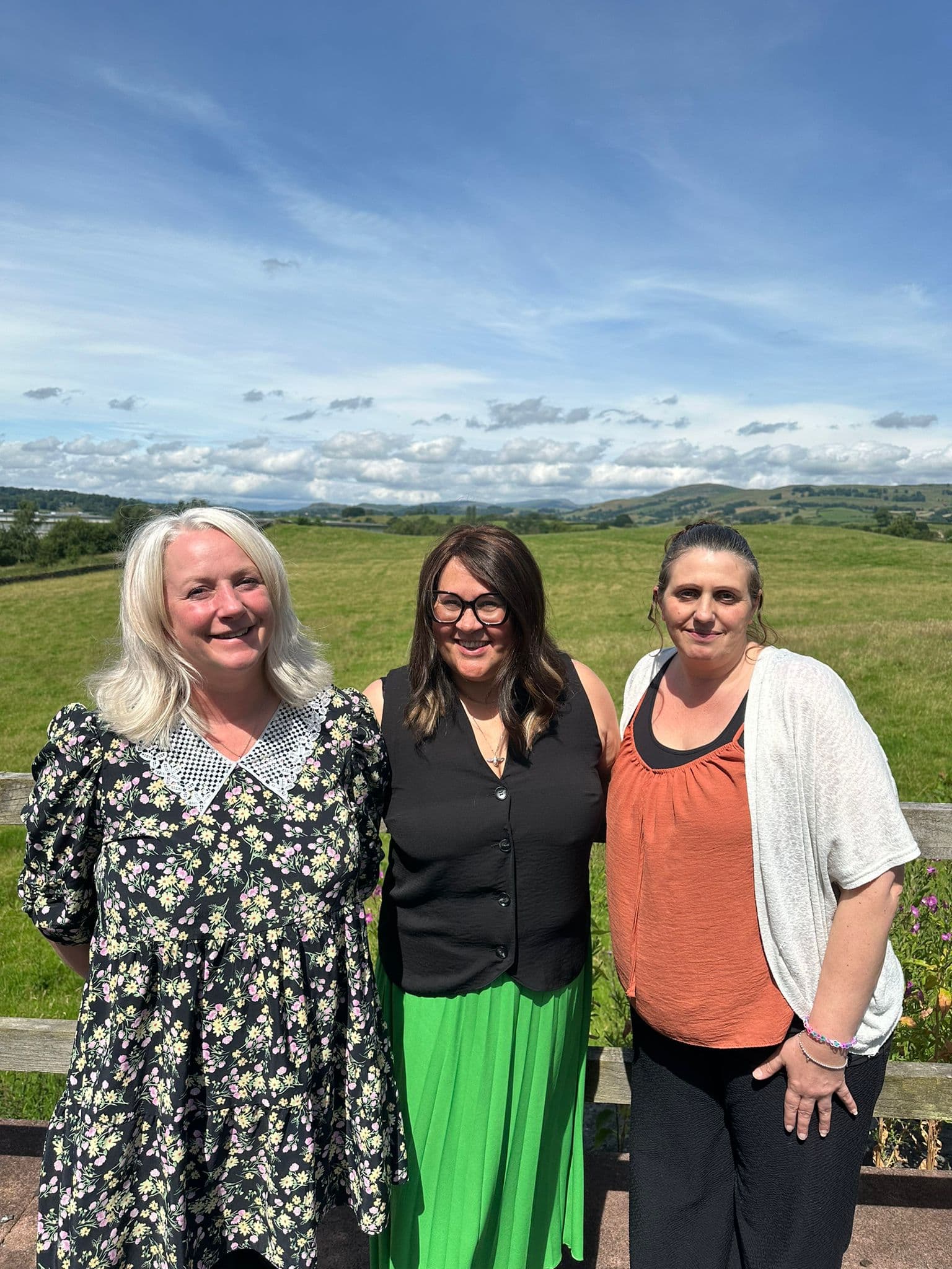 Three smiling women standing in front of a scenic landscape with green fields and a blue sky. - Home Instead