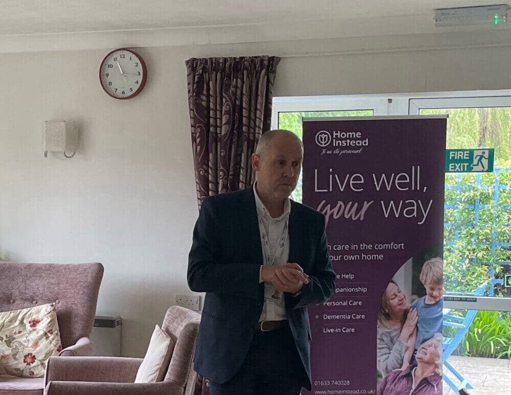 A man in a suit stands near a "Home Instead" banner, speaking in a room with a wall clock and a fire exit sign. - Home Instead