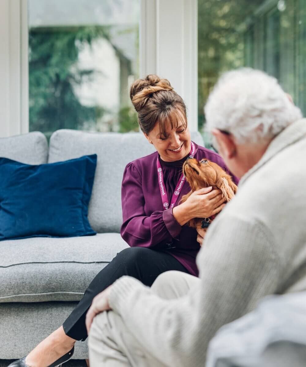 Woman and elderly man sitting on a couch, woman holding and petting a small dog. - Home Instead