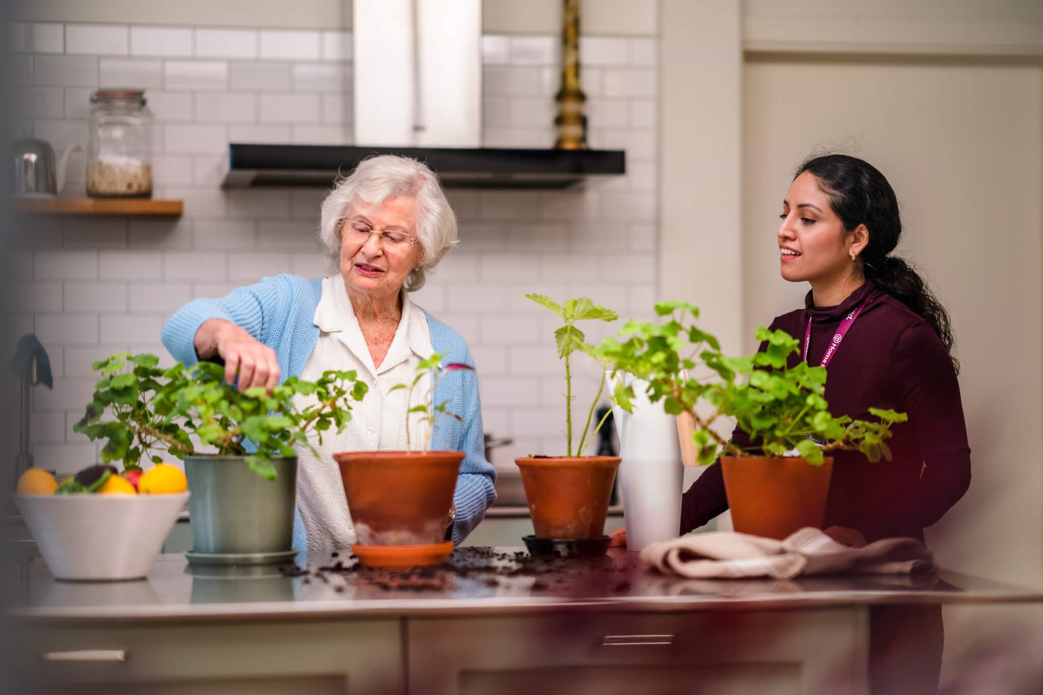 Older woman and younger woman tending to potted plants on a kitchen counter, with soil and pots around them. - Home Instead