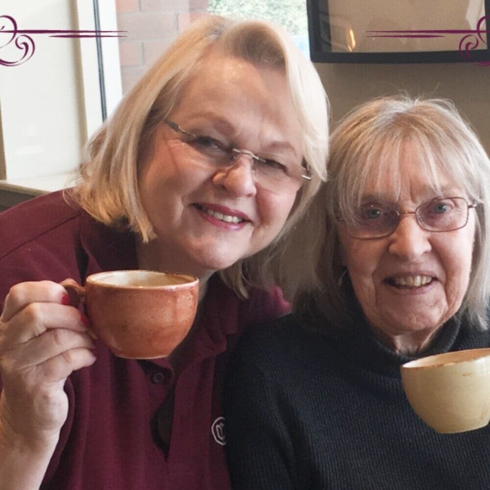 Two elderly women smiling and holding coffee cups, sitting closely together in a cozy environment. - Home Instead