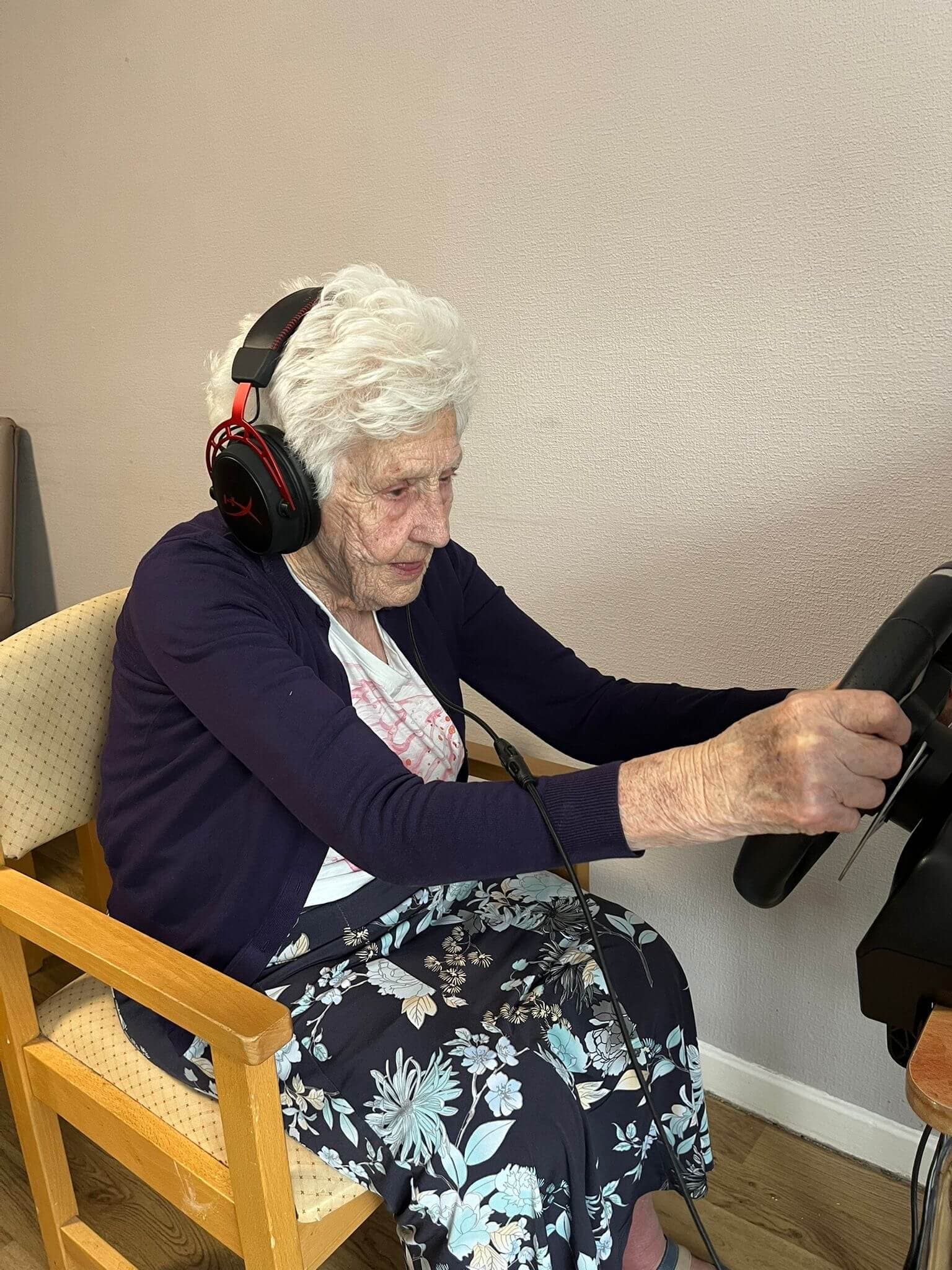 Elderly woman wearing headphones plays with a racing simulator, steering wheel in hand, seated in a floral skirt and sweater. - Home Instead