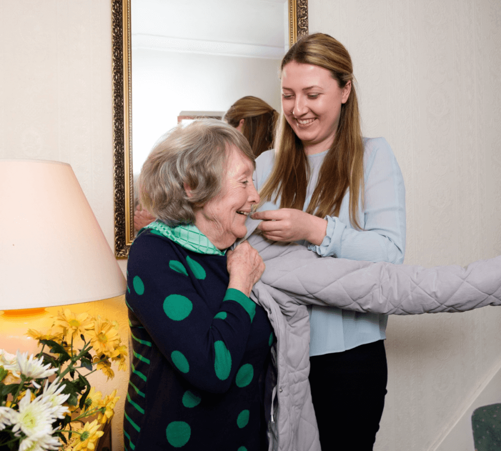Young woman helps older woman put on a coat near a mirror, both smiling; flowers and lamp in the background. - Home Instead