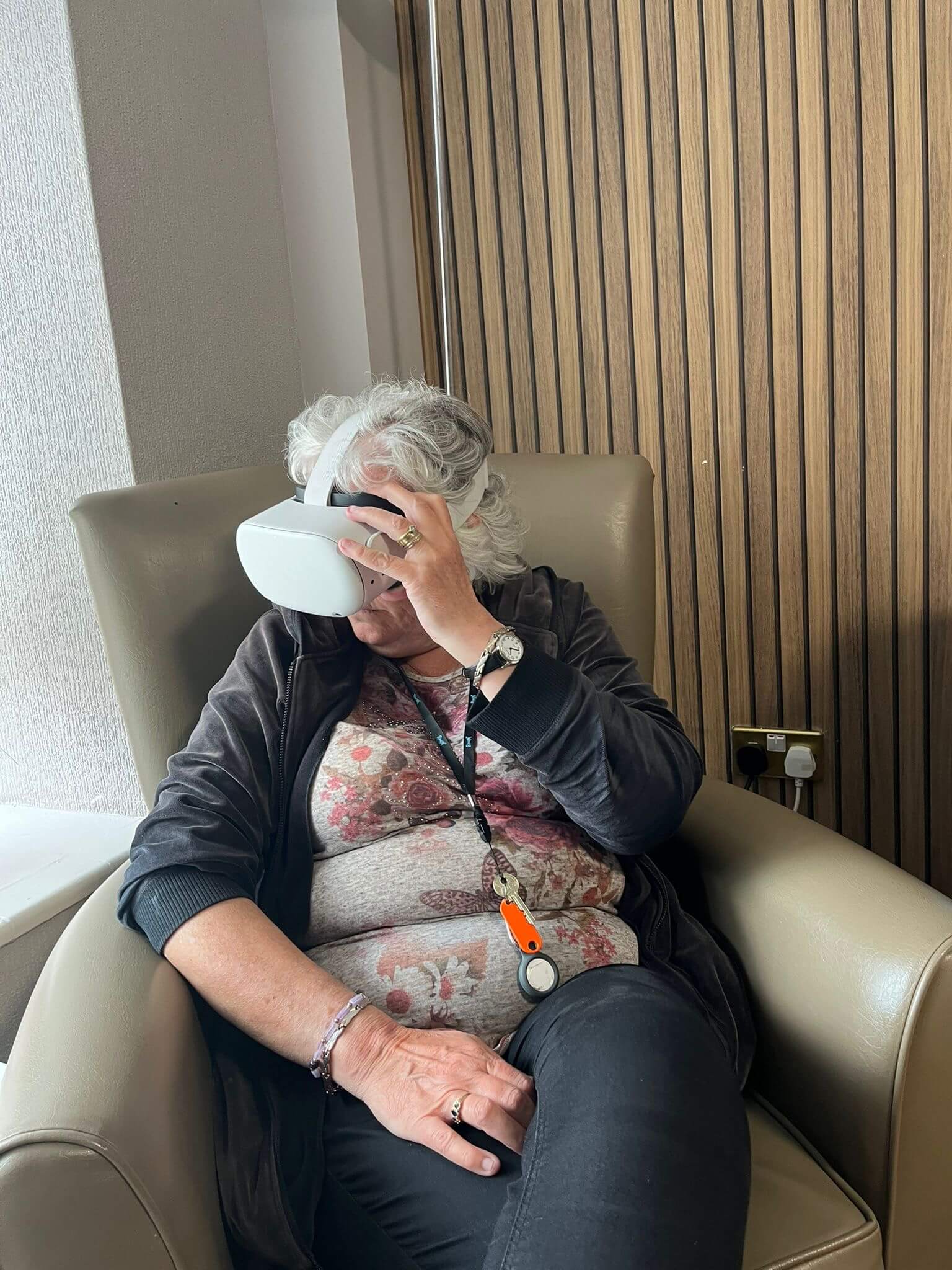 An elderly woman sits in an armchair wearing a VR headset, partially covering her face with her hand. - Home Instead