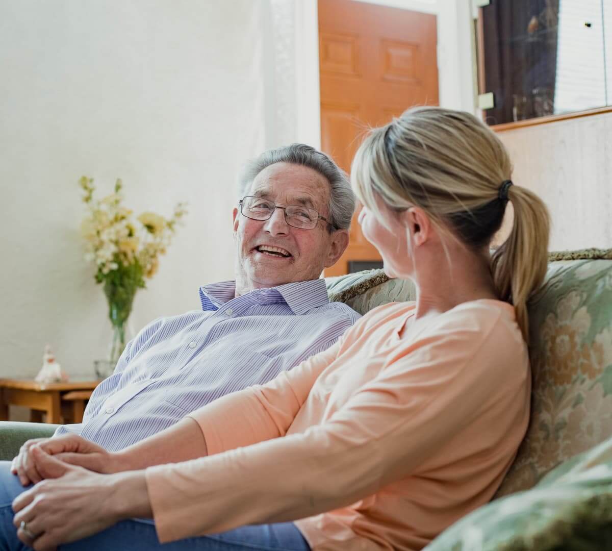 An elderly man and a younger woman sit on a couch, smiling and holding hands in a well-lit living room. - Home Instead