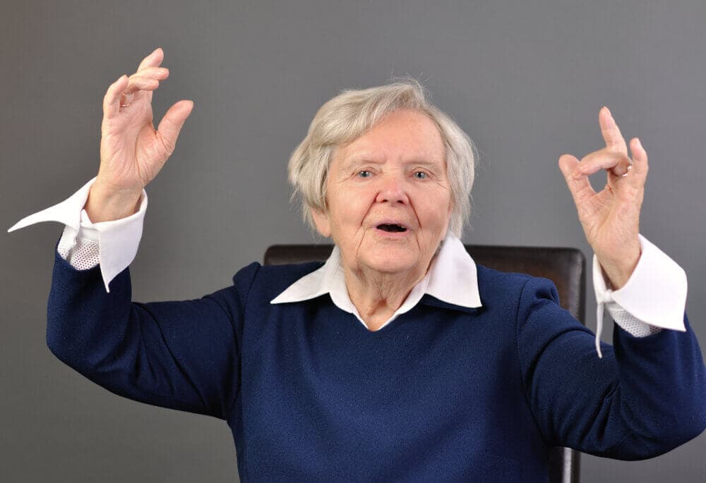 Older woman with short gray hair, wearing a navy blue sweater, gestures expressively with both hands in front of a gray background. - Home Instead