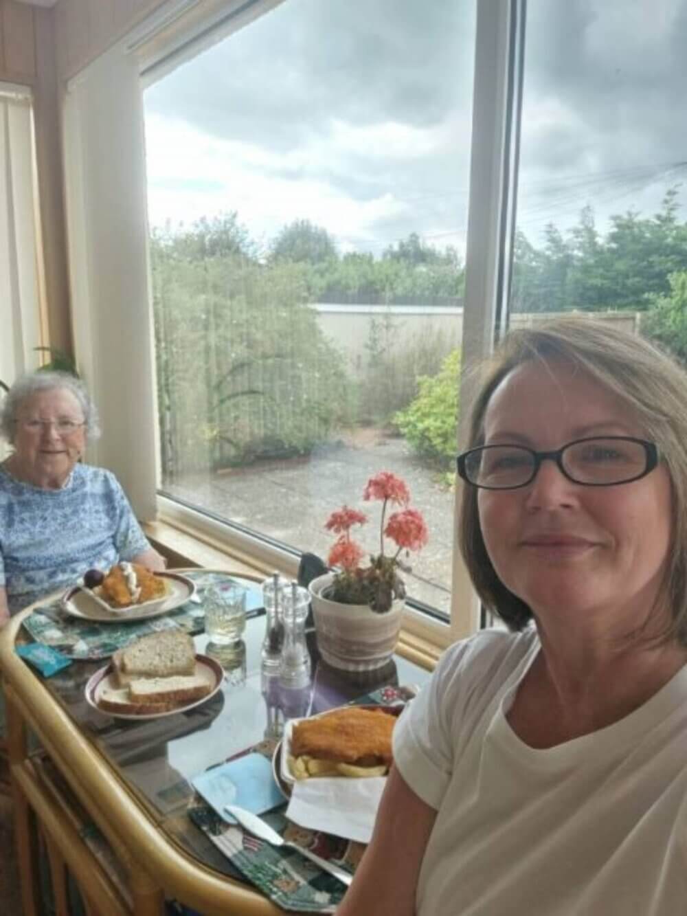 Two women enjoying a meal at a table by a large window with a garden view. - Home Instead