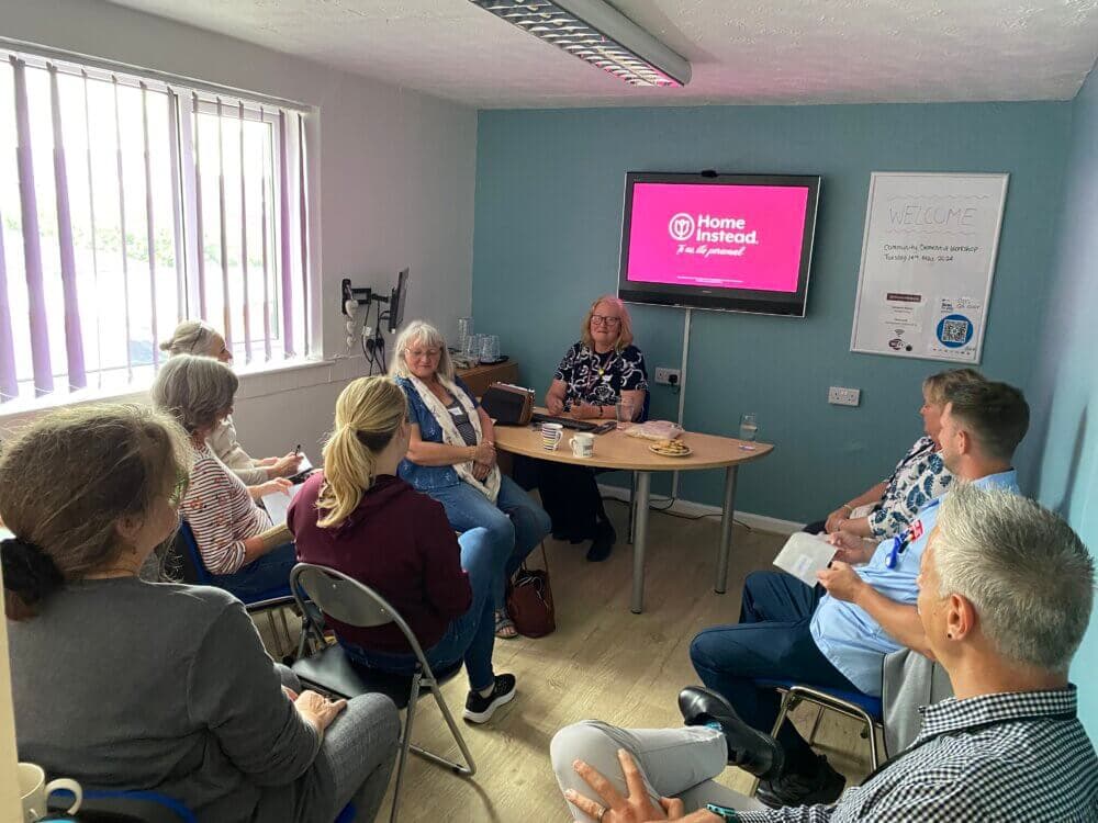 A group of people sit in a room, having a meeting. A screen displays a "Home Instead" presentation. - Home Instead