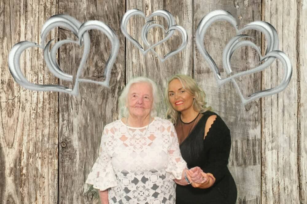 Two women, one elderly and one middle-aged, stand in front of a wooden backdrop with silver heart decorations. - Home Instead