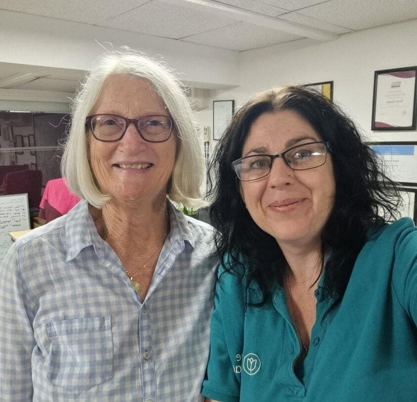 Two women with glasses smiling at the camera in an office setting. One wears a blue checkered shirt, the other a teal shirt. - Home Instead