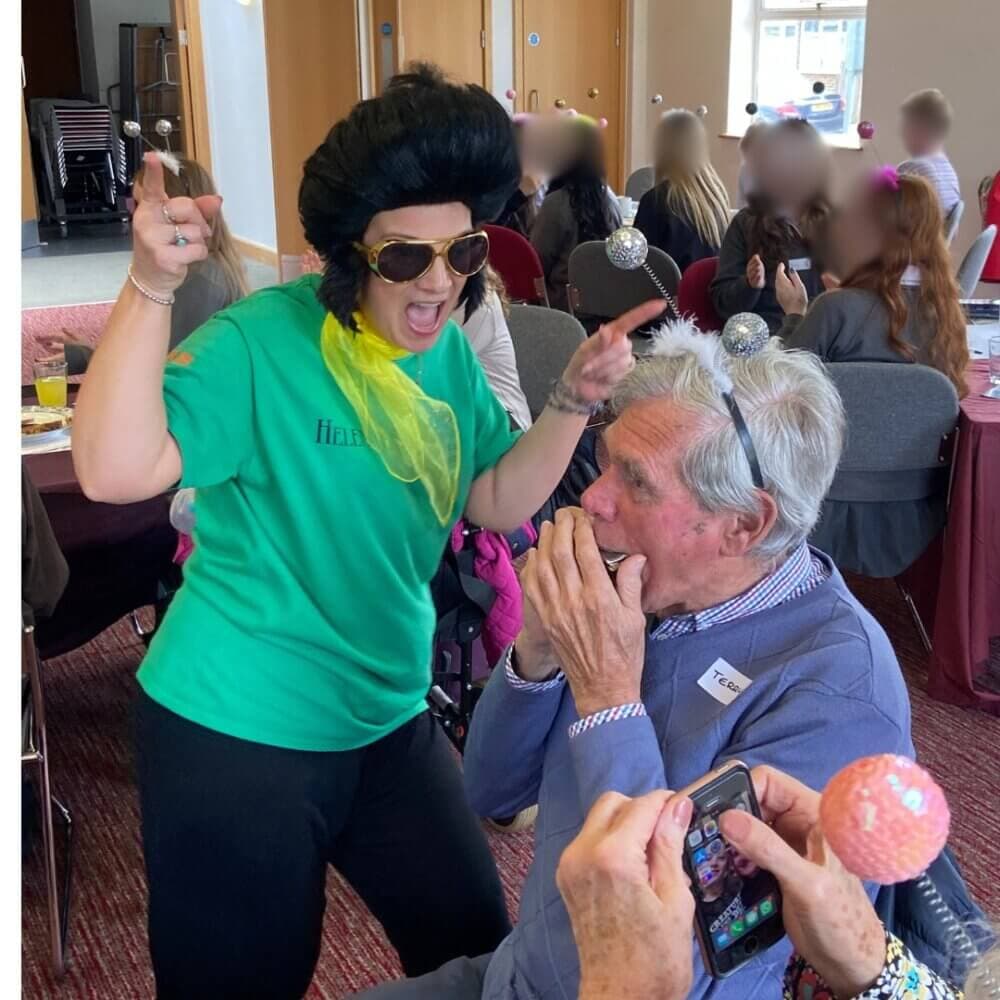Person dressed as Elvis Presley singing at Home Instead event and elderly main playing the harmonica