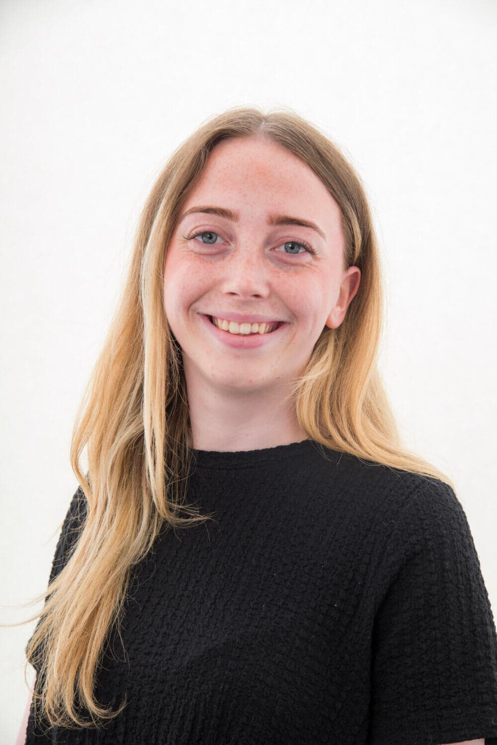 A young woman with long blonde hair smiles at the camera, wearing a black textured top against a plain white background. - Home Instead
