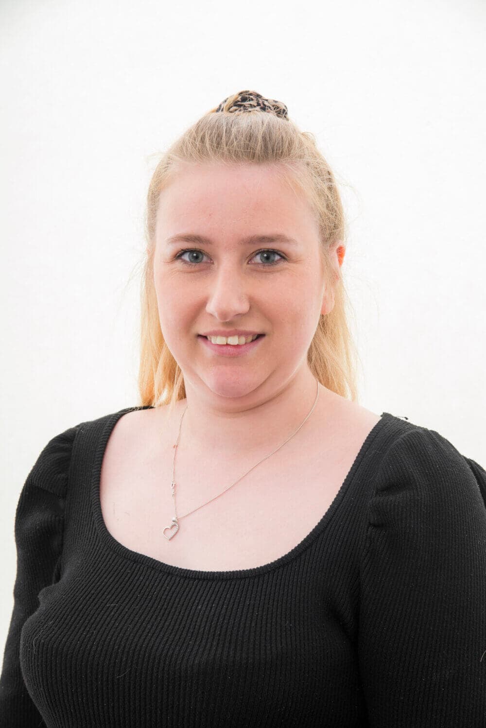 A woman with long blonde hair, wearing a black top and a necklace, smiles at the camera against a white background. - Home Instead