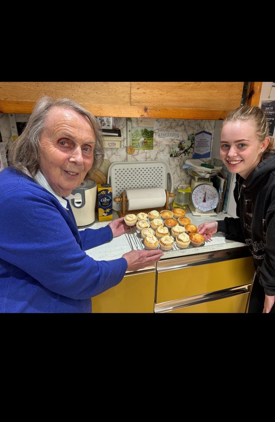 Two women smiling, holding a tray of freshly baked muffins in a kitchen. - Home Instead