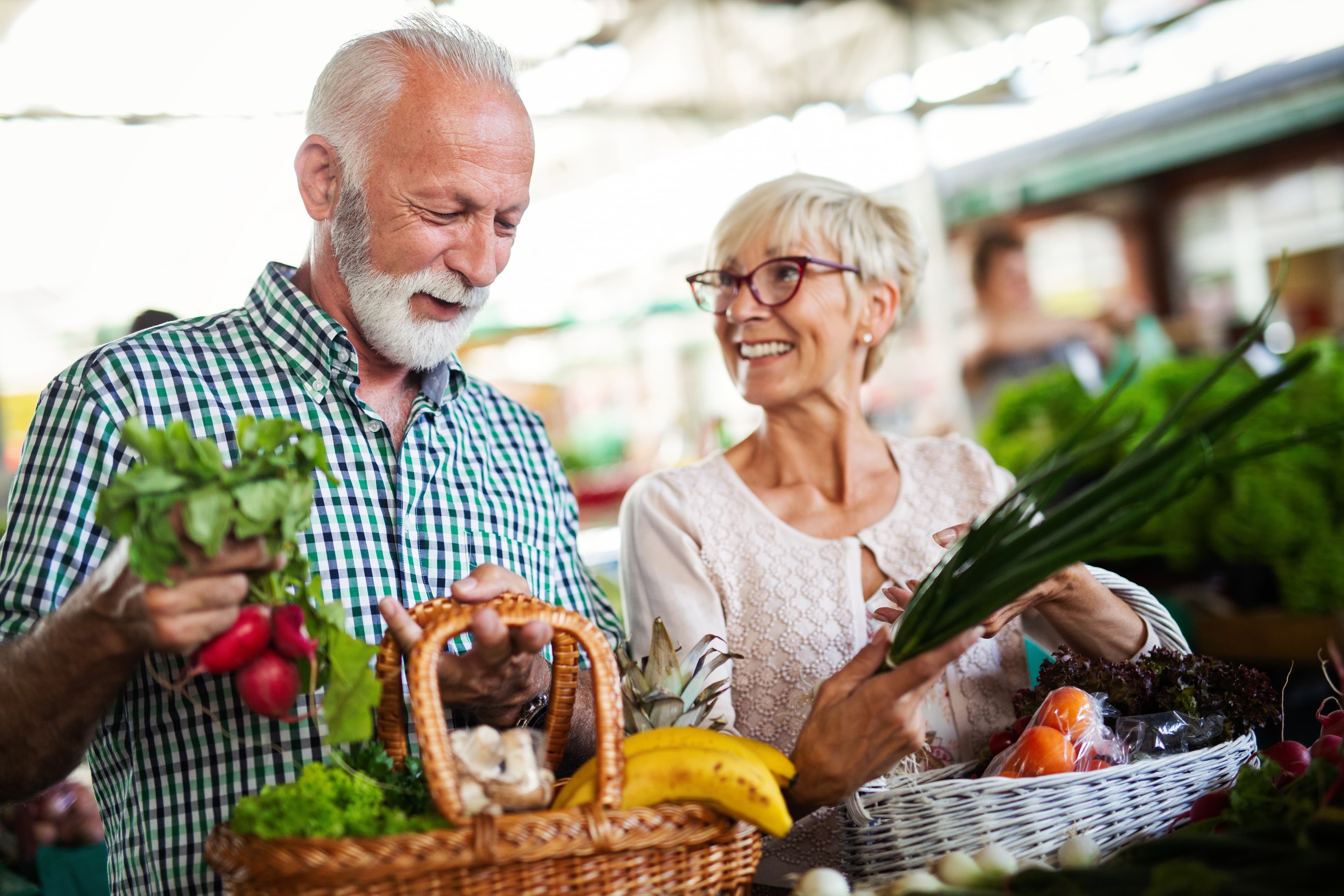 An elderly man and woman smile while holding baskets full of fresh vegetables at an outdoor market. - Home Instead