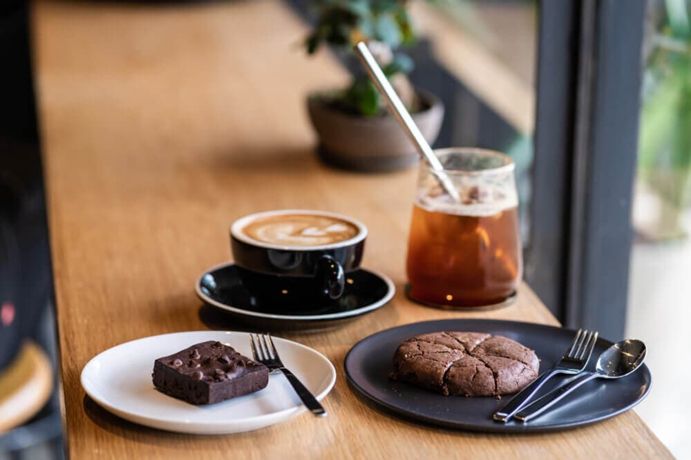 A coffee and iced tea with a brownie on a white plate and a cookie on a black plate, set on a wooden table. - Home Instead