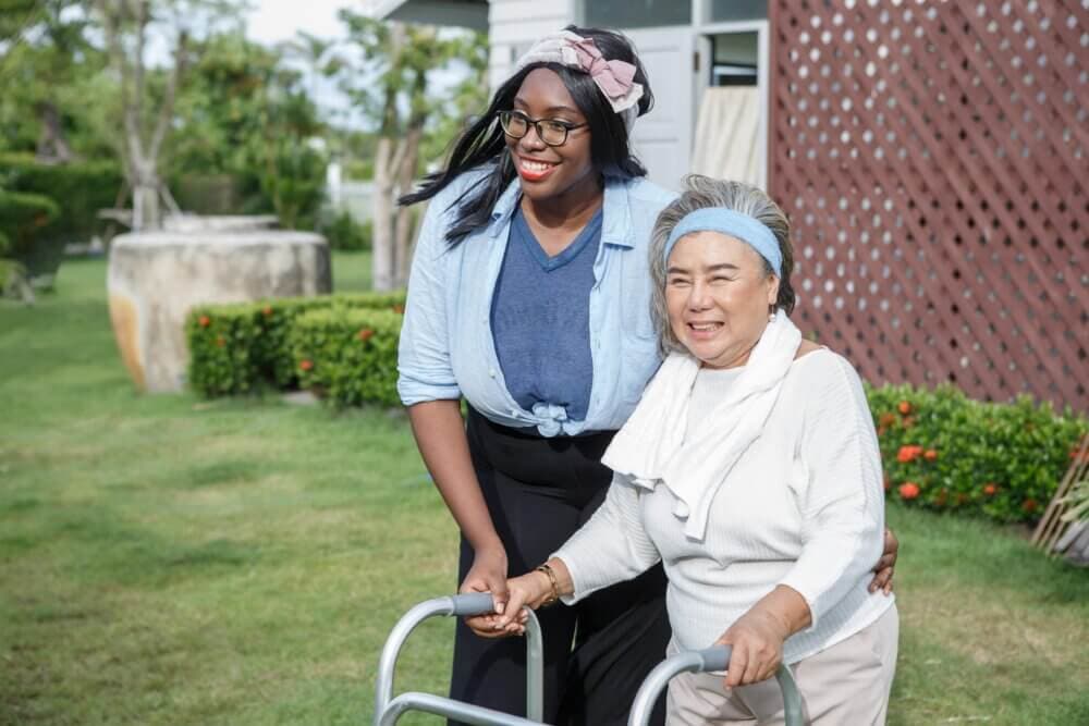 A woman helps an elderly woman with a walker outside on a sunny day, both smiling. - Home Instead