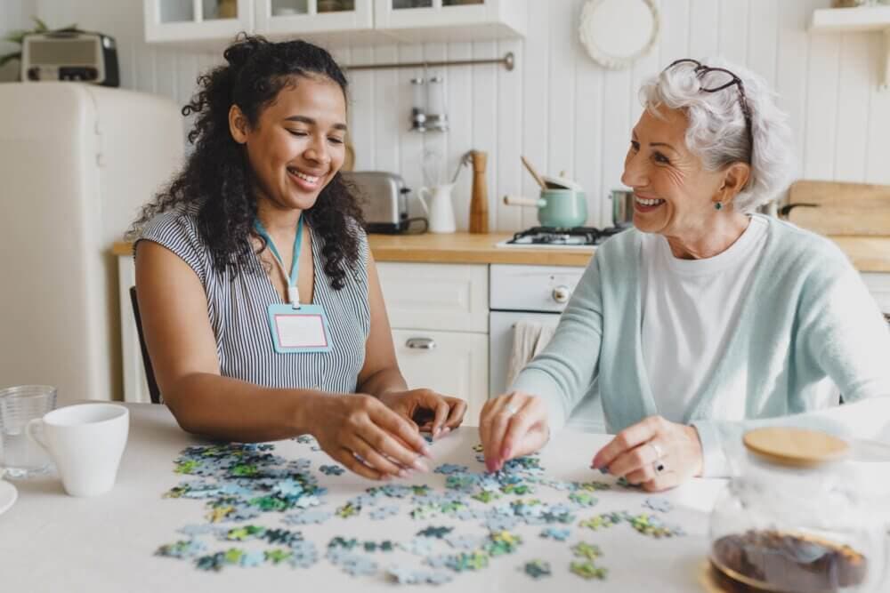 Two women, one younger and one older, smiling and working on a jigsaw puzzle together at a kitchen table. - Home Instead