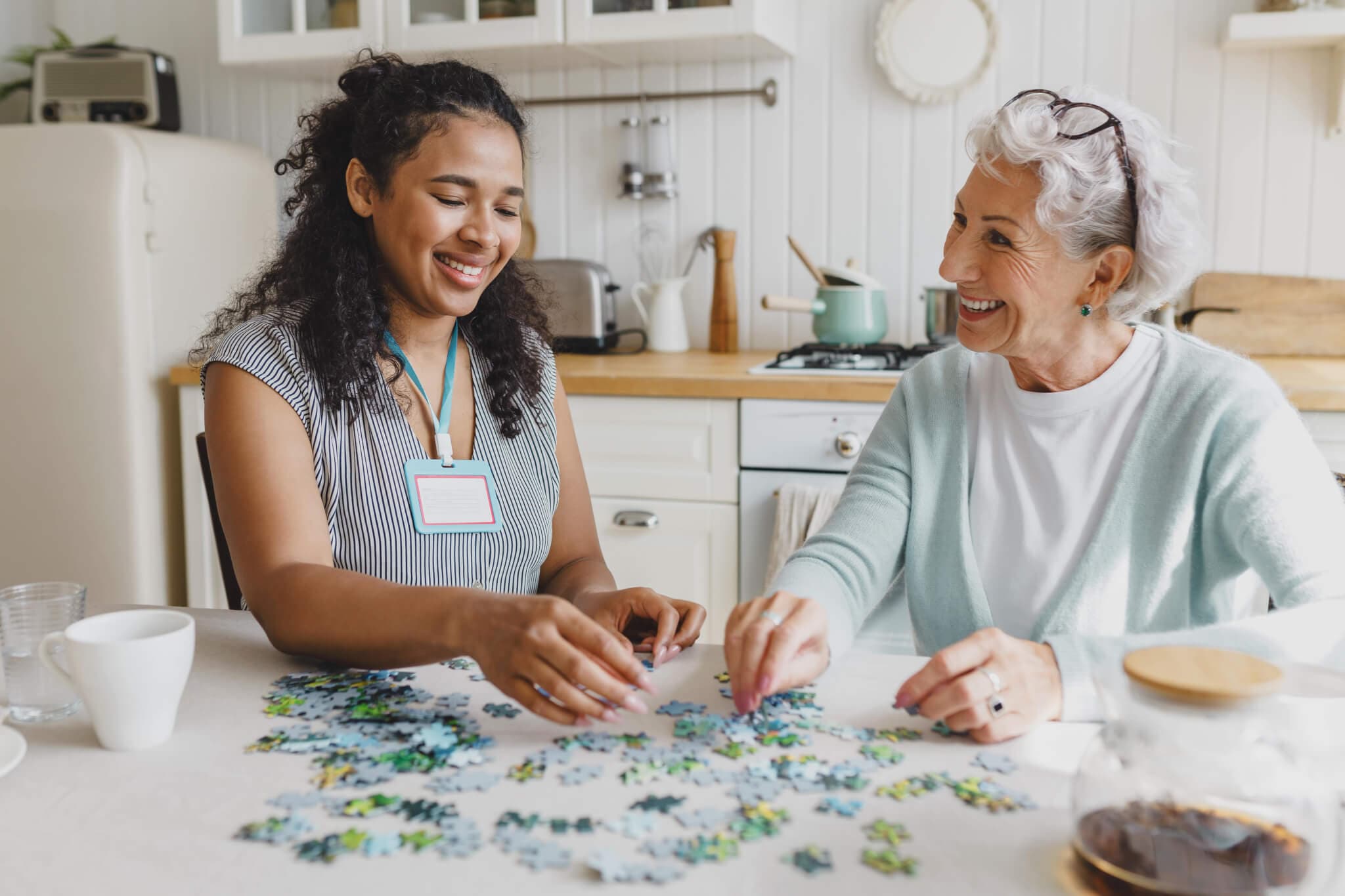 Two women, one younger and one older, smiling and working on a jigsaw puzzle together at a kitchen table. - Home Instead