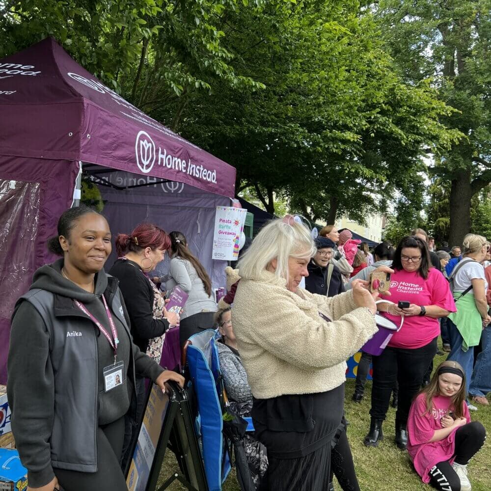 People gathered at an outdoor community event, with a purple Home Instead tent and various activities happening. - Home Instead