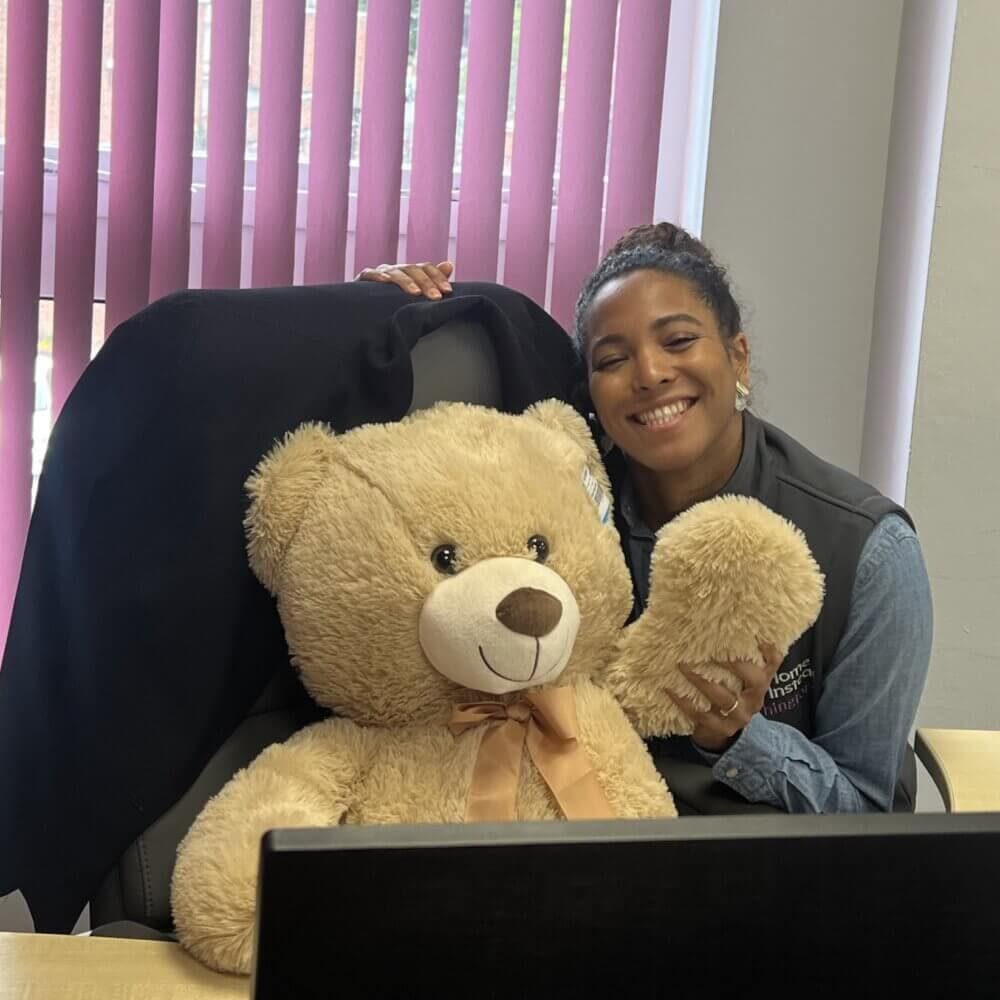 A smiling woman poses with a large teddy bear sitting on an office chair behind a desk with a computer. - Home Instead