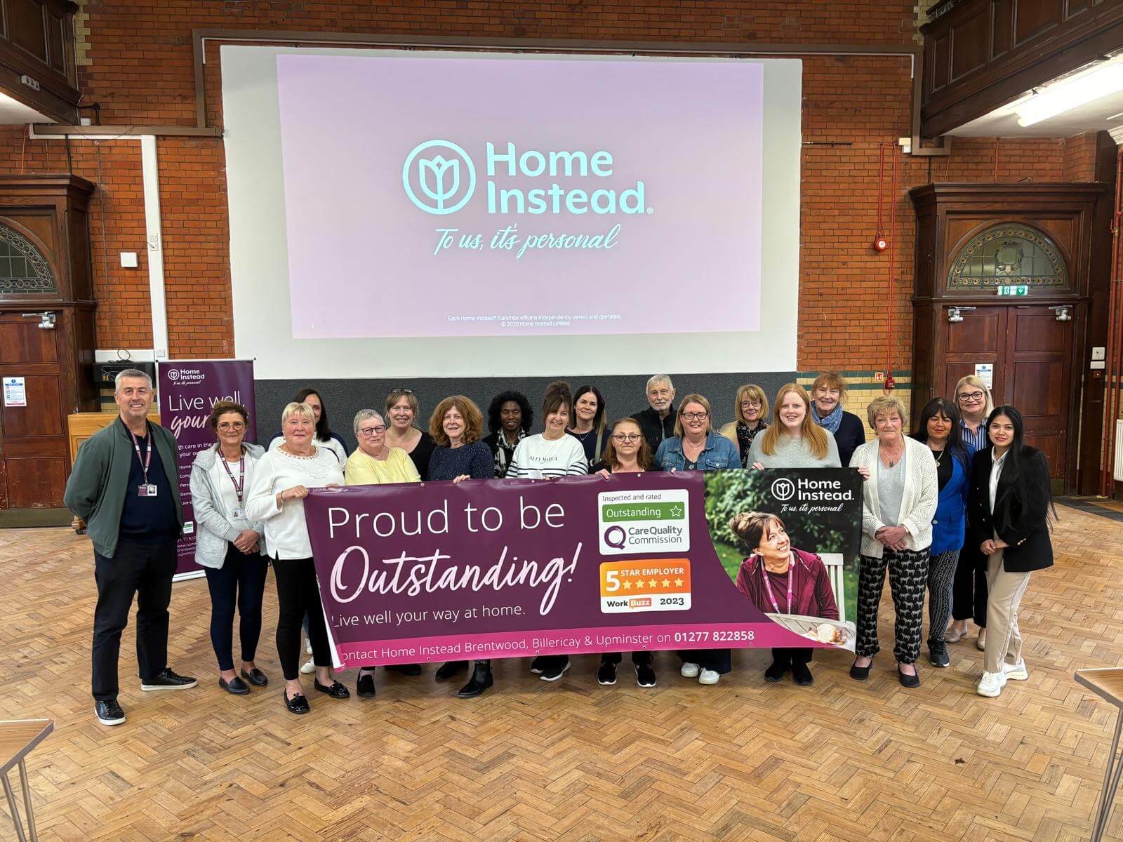 A group of people standing in front of a screen and holding a "Proud to be Outstanding!" banner indoors. - Home Instead