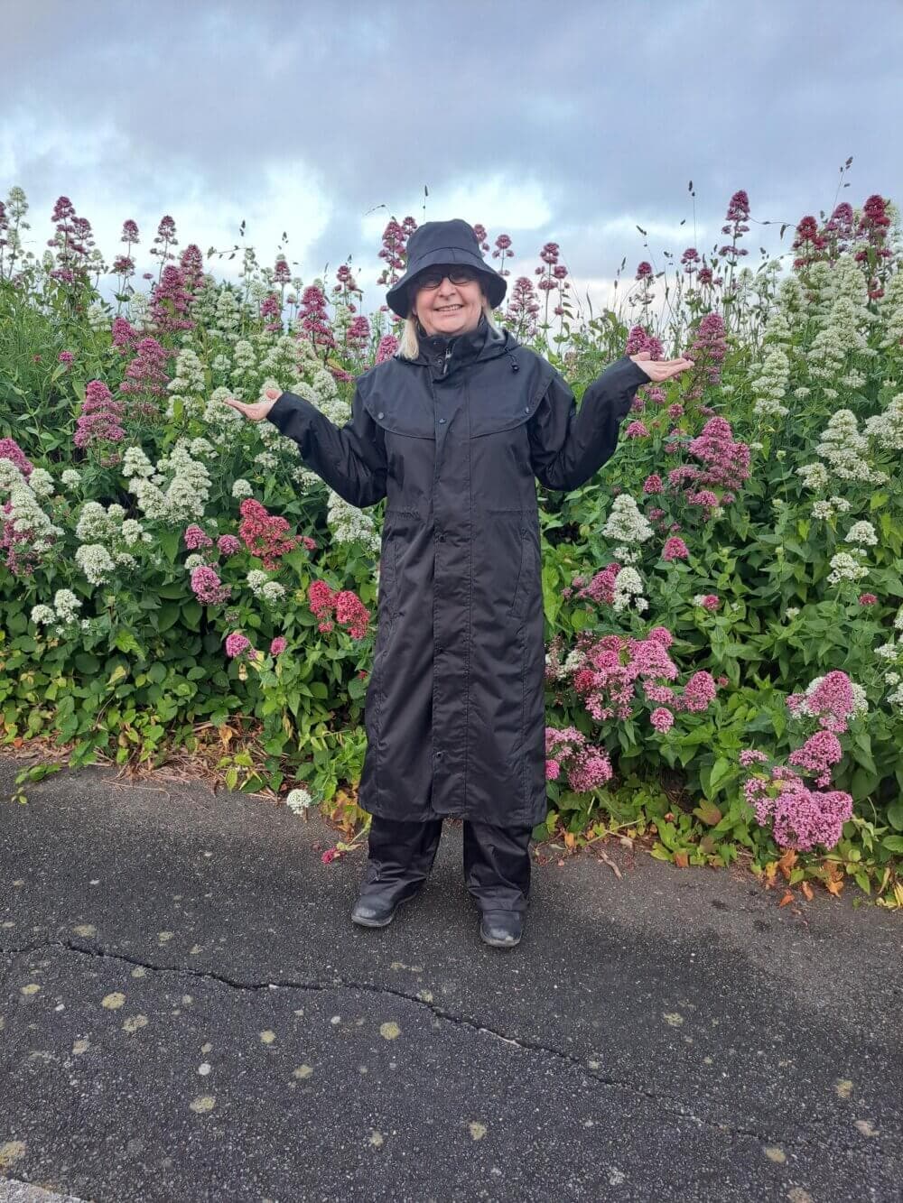 Person in a black raincoat and hat stands on a paved path, smiling with arms outstretched, in front of colorful flowers. - Home Instead